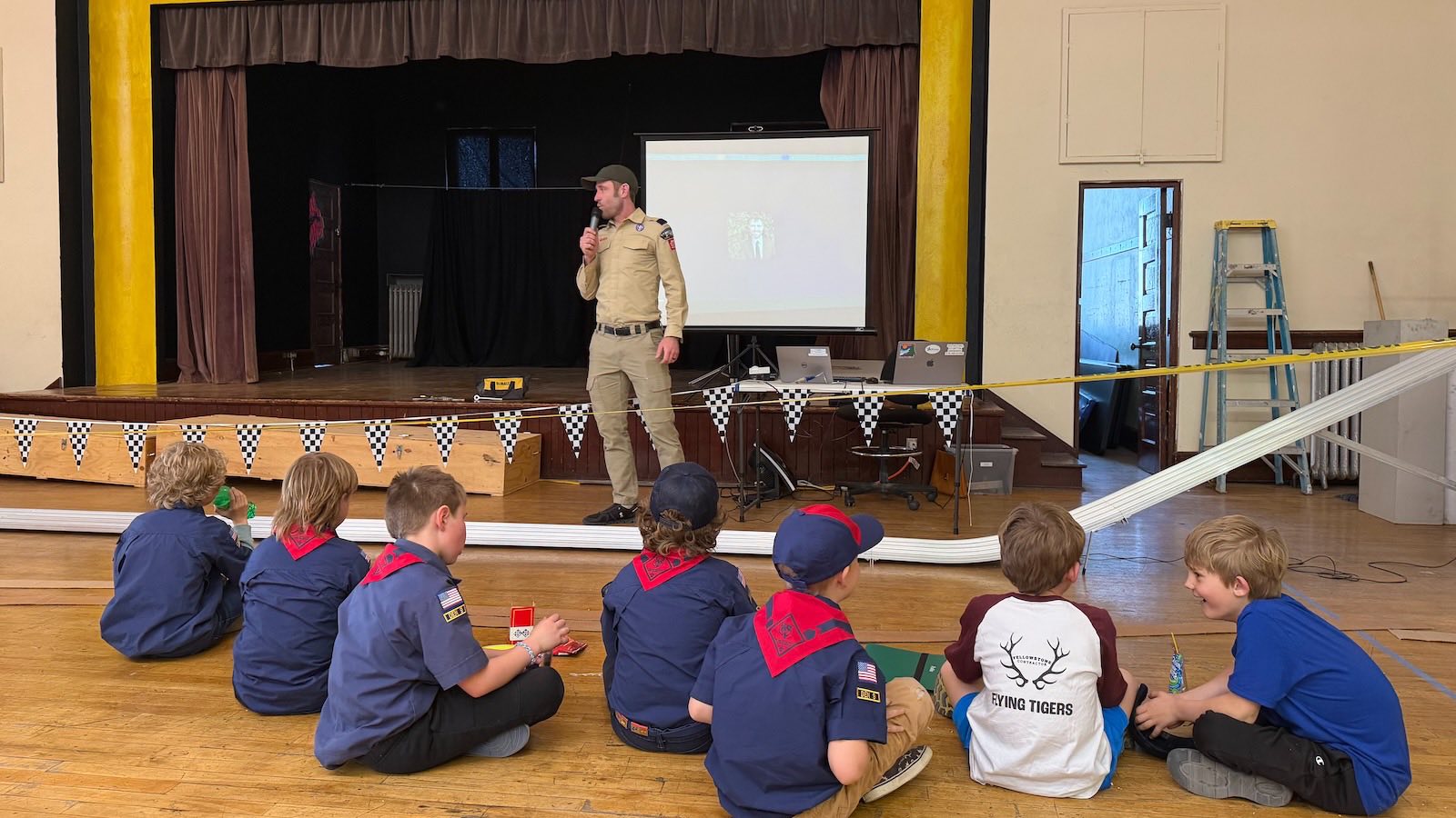 A uniformed pack leader with a microphone talking to Cub Scouts sitting on the floor beside the Pinewood Derby track